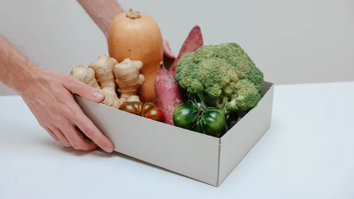 Deux mains soulèvent un carton blanc rempli de légumes frais, dont un brocoli et une courge, prêts à être expédiés suite à une commande sur une boutique en ligne pour producteur dans le Tarn.