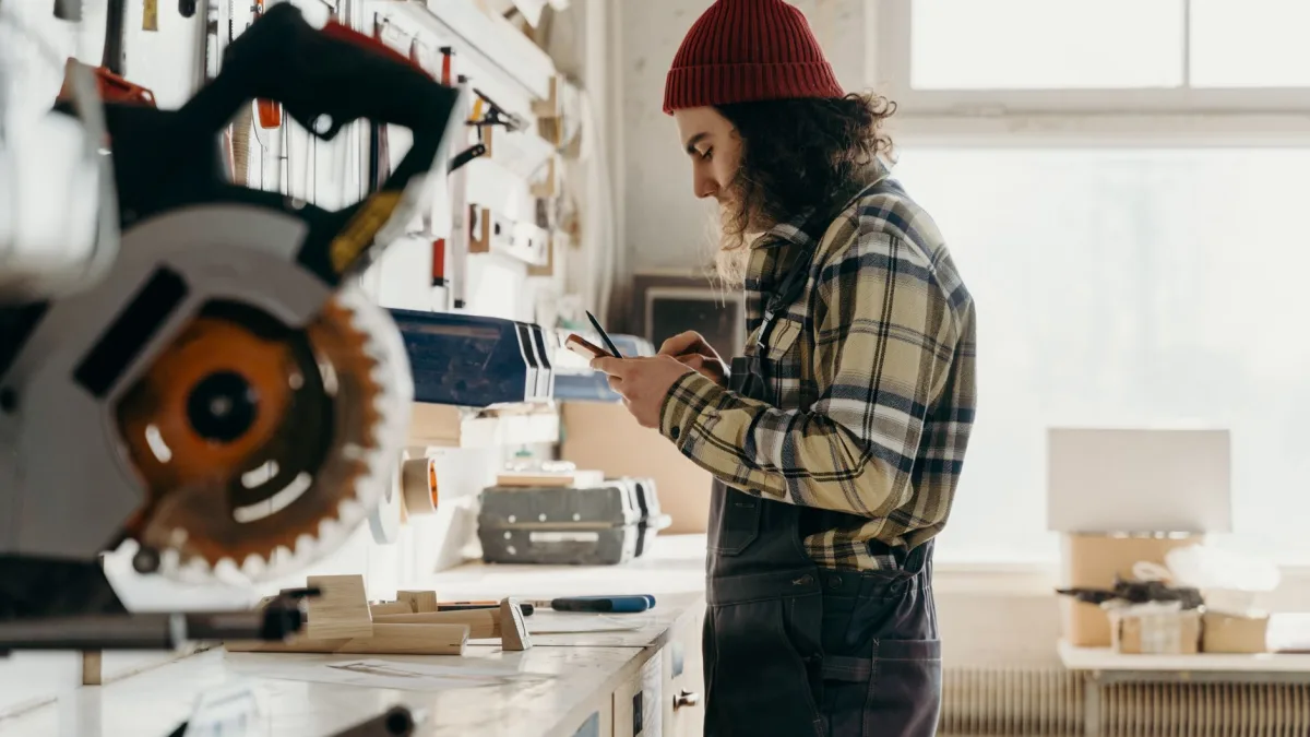 Un artisan en salopette consulte son smartphone dans un atelier de menuiserie pour utiliser son application de gestion des salariés à Perpignan.