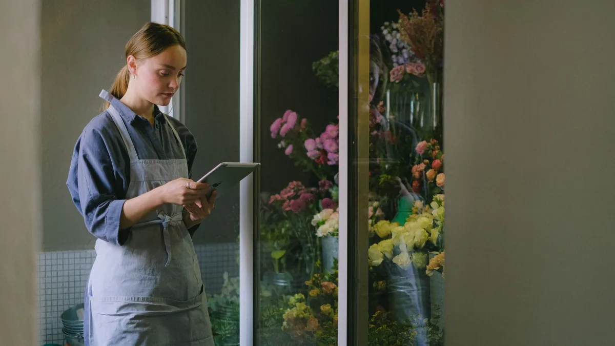 Une fleuriste en tablier utilise une tablette numérique devant une vitrine de fleurs dans sa boutique pour consulter son CRM sur mesure à Perpignan.