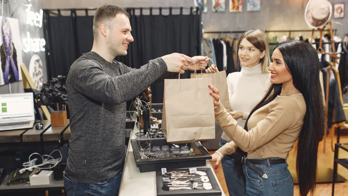 Un vendeur de boutique tend des sacs en papier à deux clientes souriantes devant un comptoir, illustrant l'utilisation d'un outil de gestion CRM dans le Tarn.